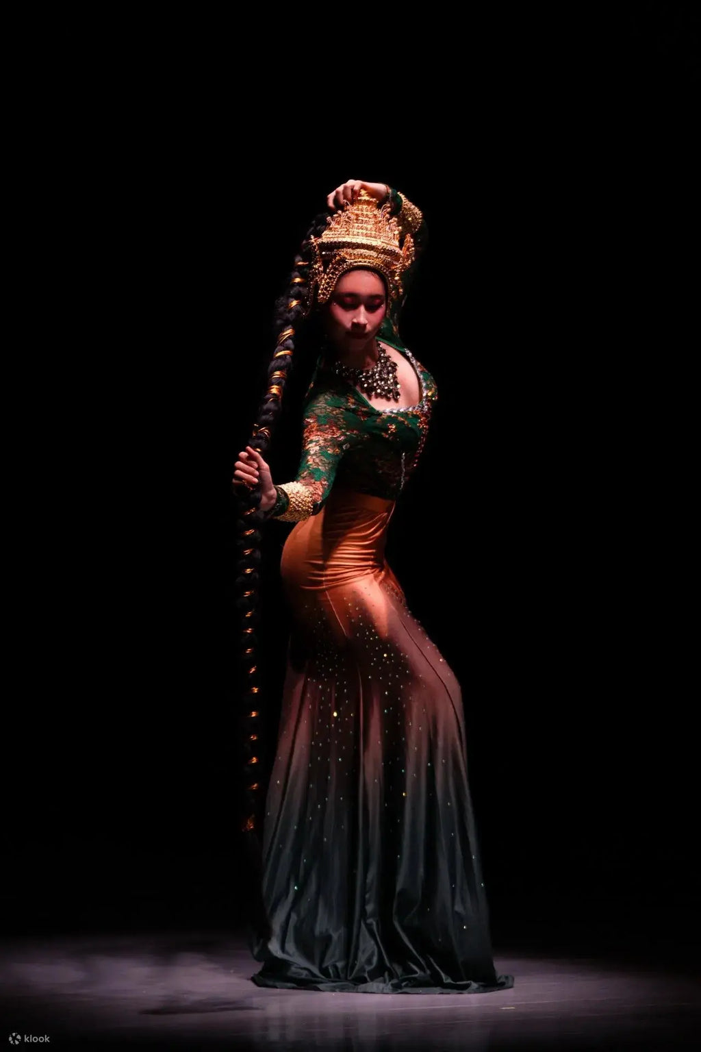 Dancer in traditional costume with intricate headpiece and long staff on a dark background